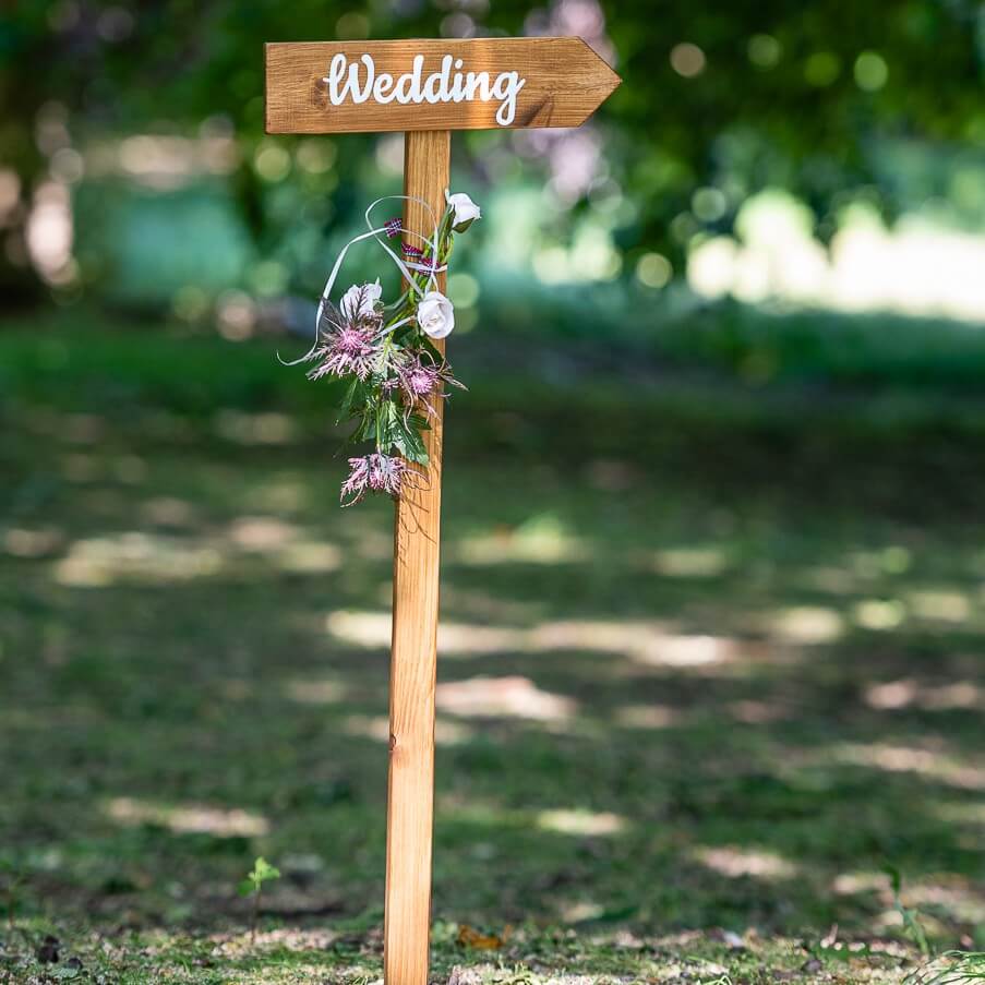 Rustic wooden directional signage for wedding ceremony, set on grass with floral details.