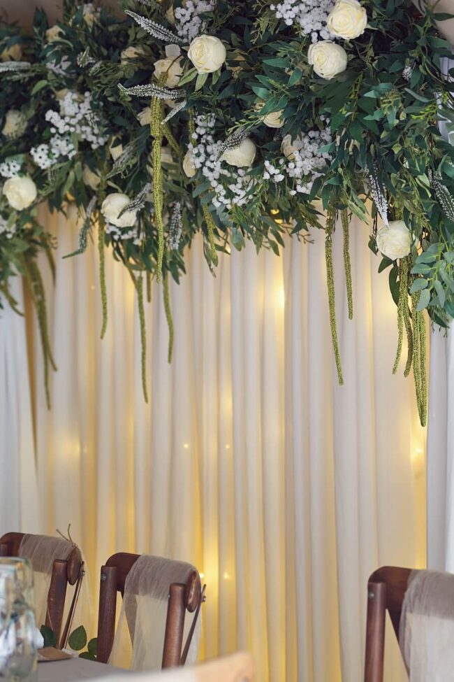Close view of a white draped wedding backdrop with warm lighting, greenery and white flowers above the top table