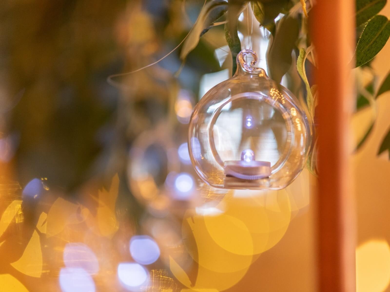 Glass hanging tealight holder suspended among leaves with warm lights behind