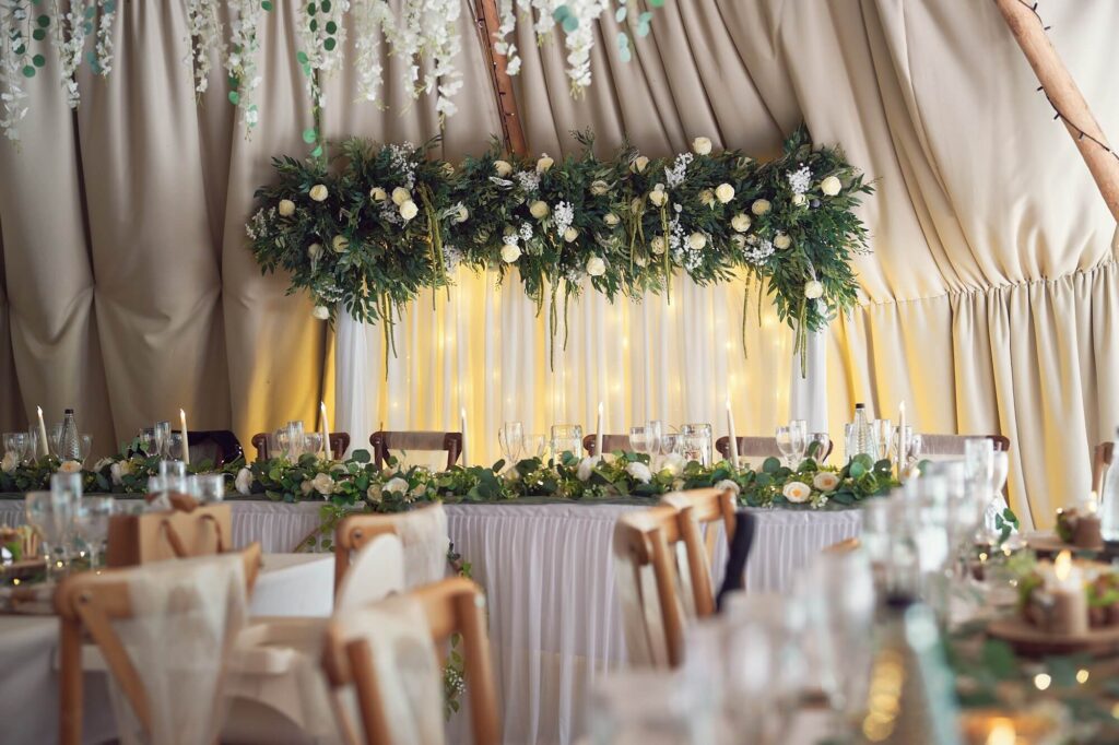 Draped floral backdrop with white fabric, warm lights, greenery and white flowers behind a wedding top table
