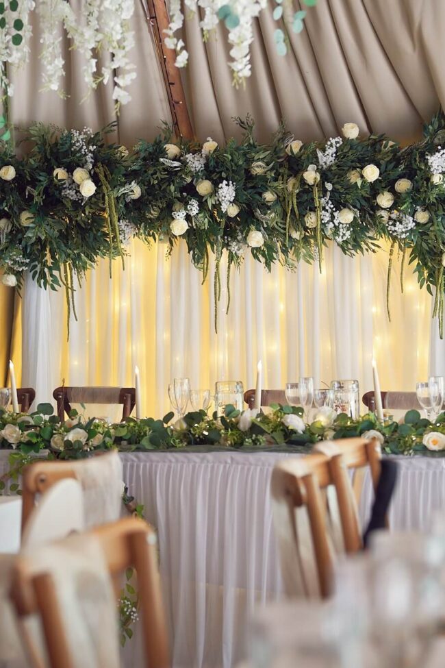 Draped floral backdrop with white fabric, warm lights, greenery and white flowers behind a wedding top table