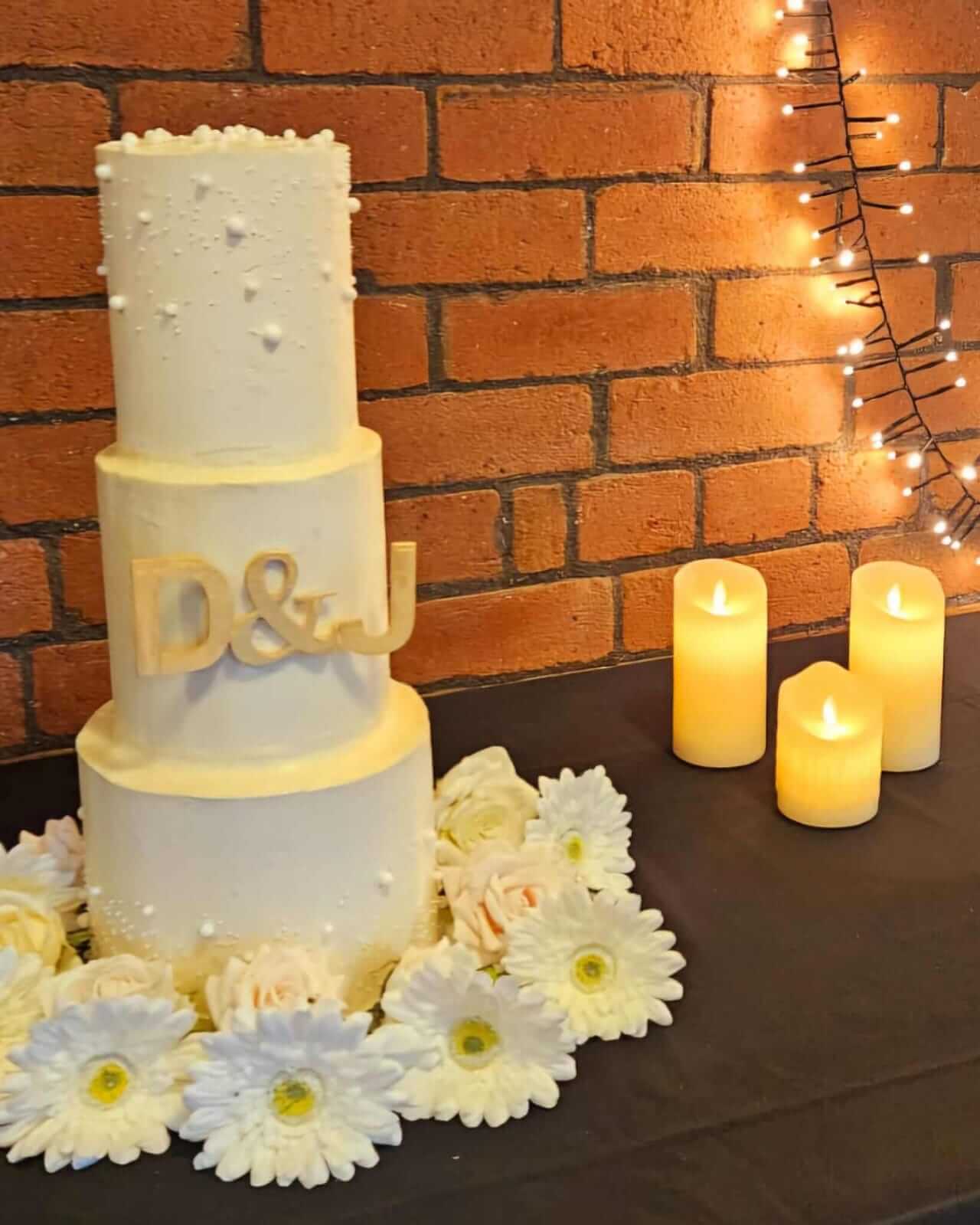 Rustic wedding cake with white flowers and candles against a brick wall.