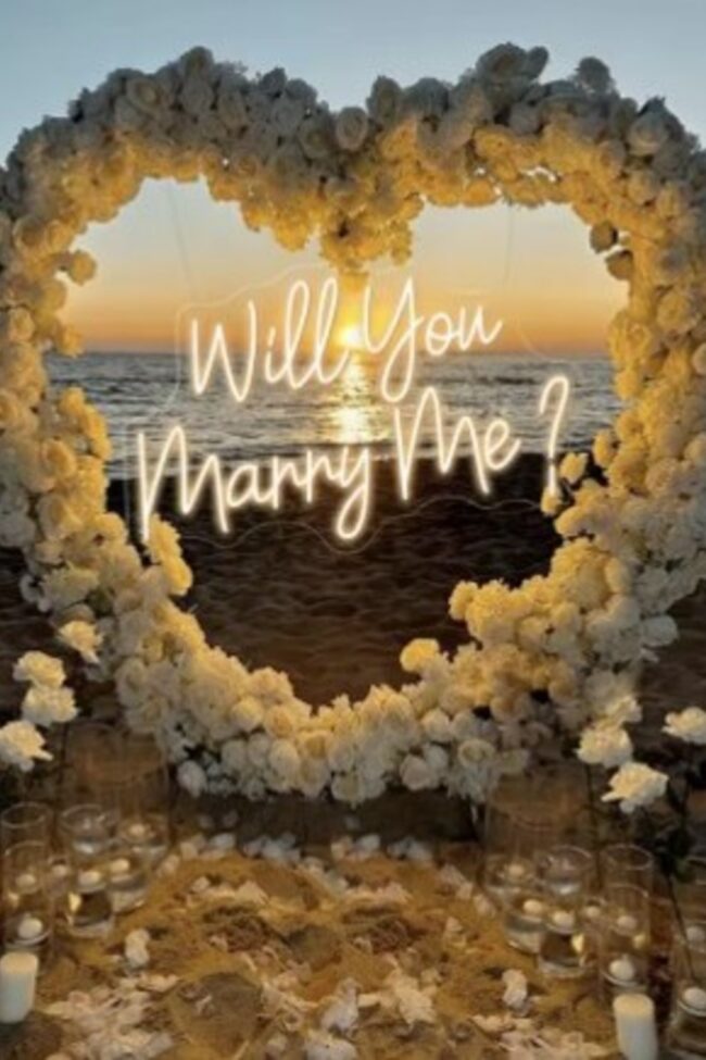 will you marry me sign lit within a floral heart arch at sunset