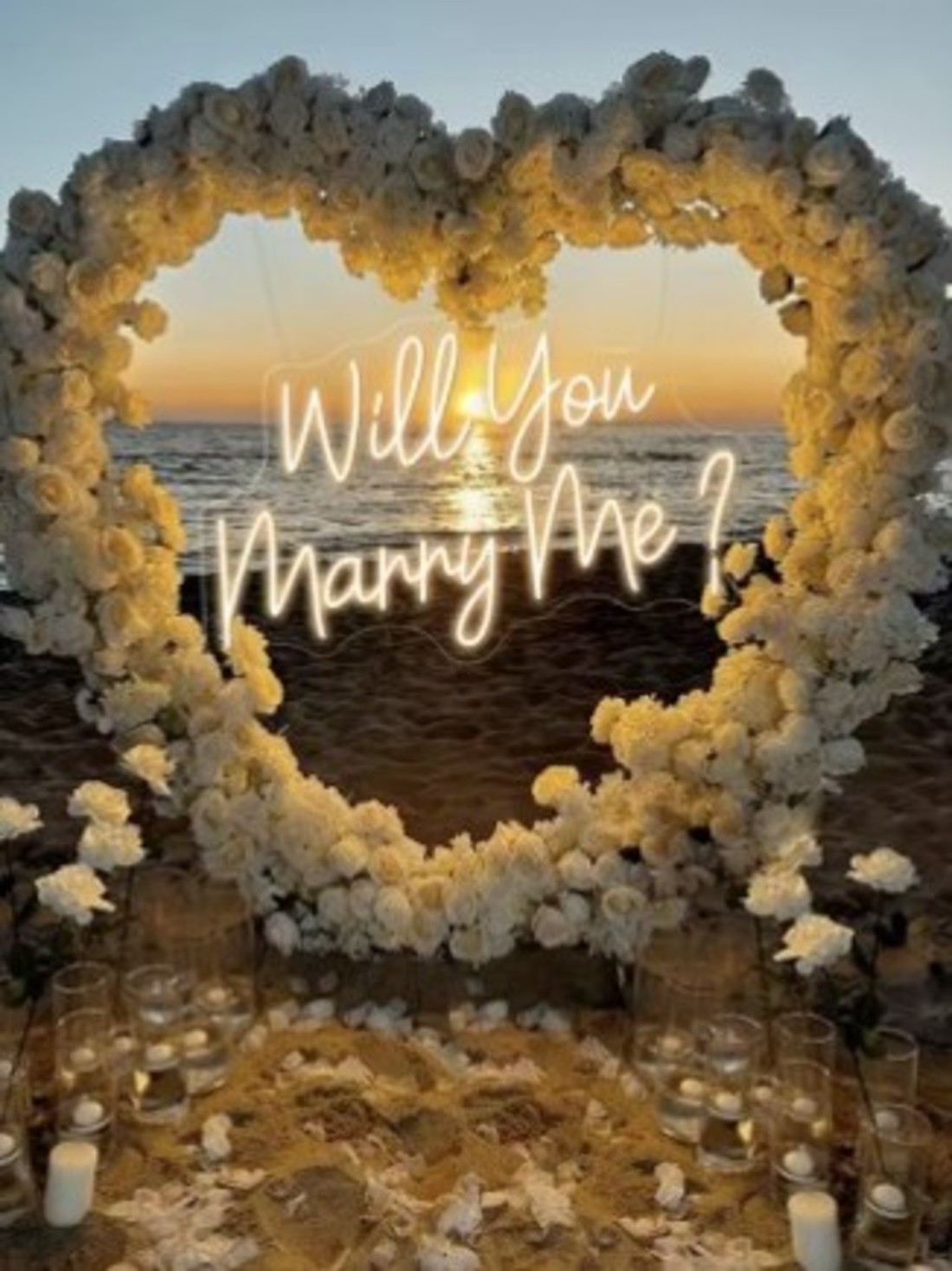will you marry me sign lit within a floral heart arch at sunset