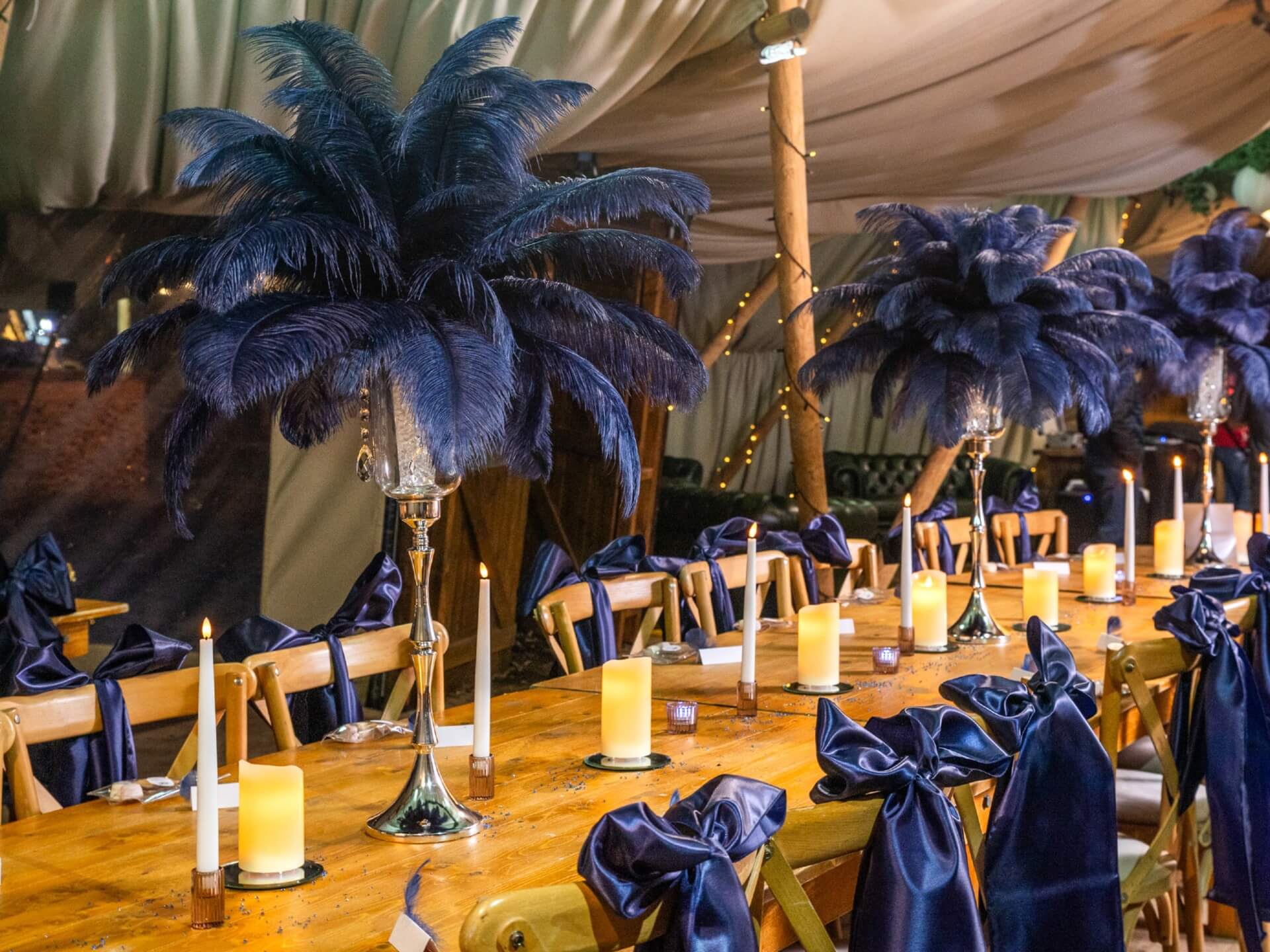 Navy blue feather centrepieces arranged along a candlelit wooden banquet table