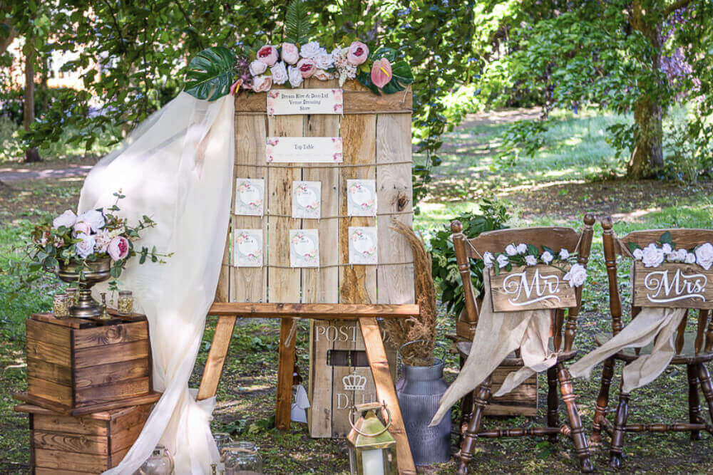 Rustic wedding stands and displays with a wooden seating plan, floral décor, and styled chairs in an outdoor garden setting.