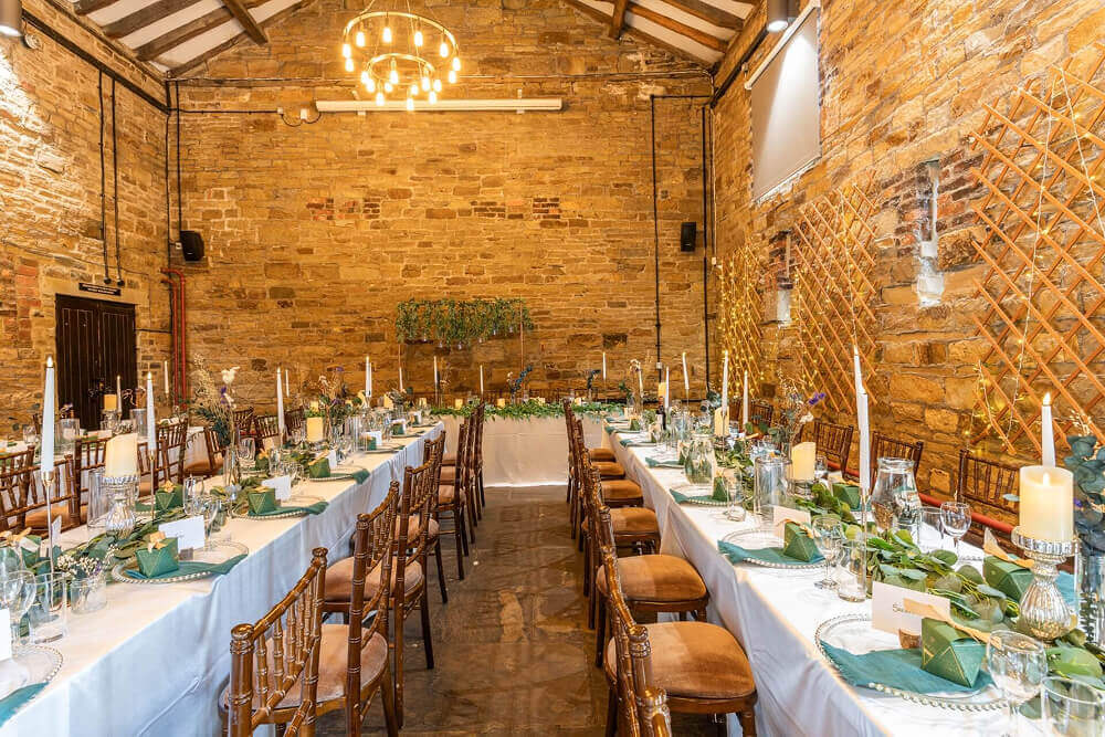 Elegant table decorations for weddings with greenery runners, candles, folded napkins and rustic wooden chairs in a stone barn venue.