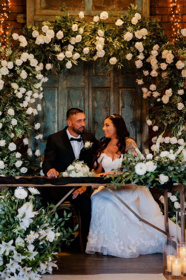 Bride and groom seated under a floral archway wedding display decorated with white roses, greenery and candles.