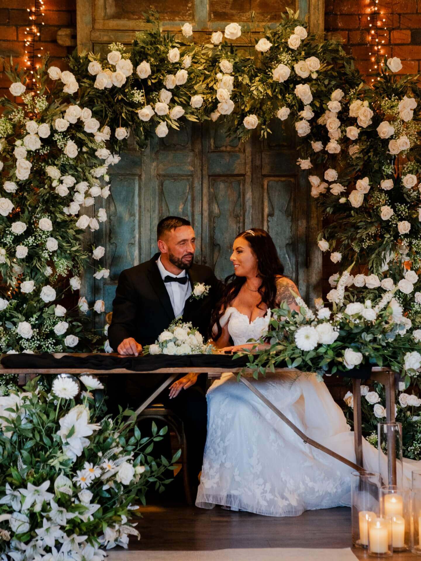 Bride and groom seated under a floral archway wedding display decorated with white roses, greenery and candles.