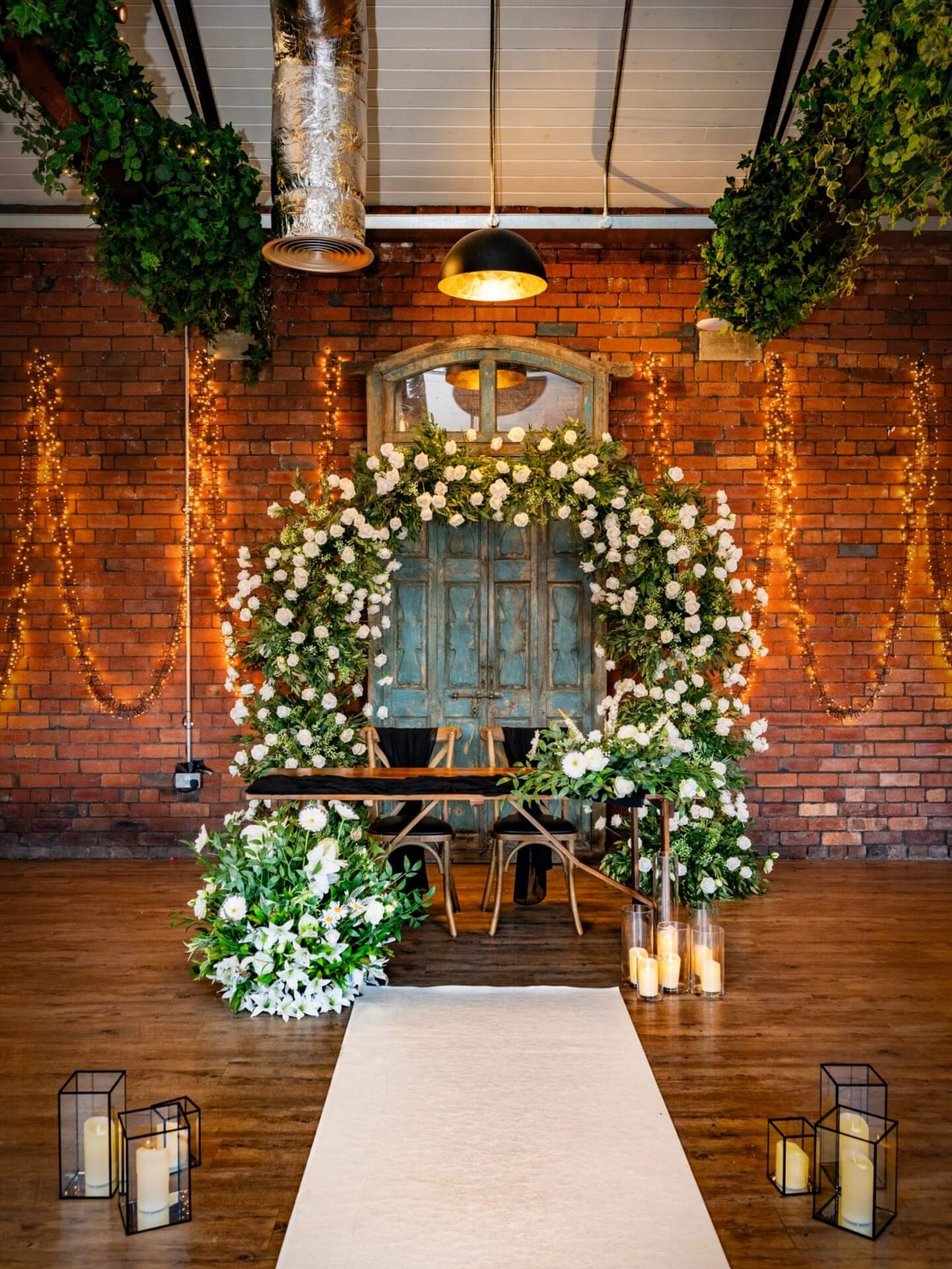 Romantic ceremony setup featuring white wedding arch flowers, greenery, candles and a rustic backdrop.
