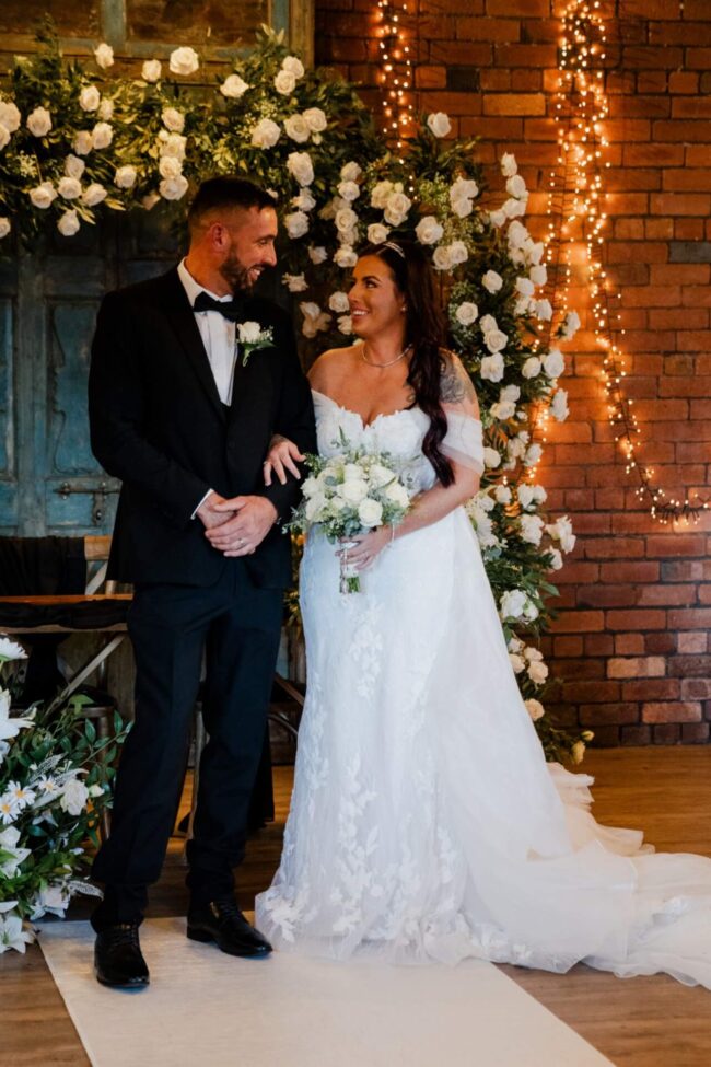 Bride and groom standing beneath a white floral arch wedding display decorated with greenery and twinkling lights.