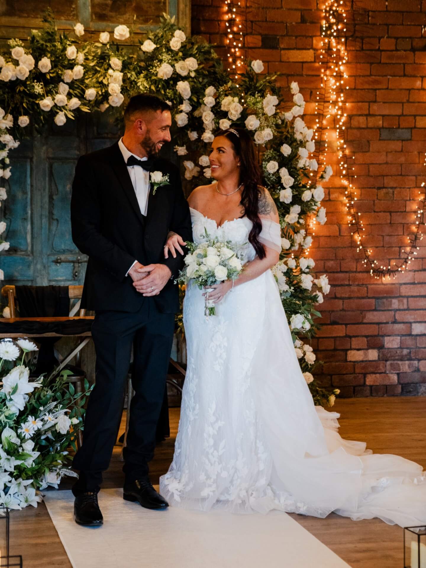 Bride and groom standing beneath a white floral arch wedding display decorated with greenery and twinkling lights.