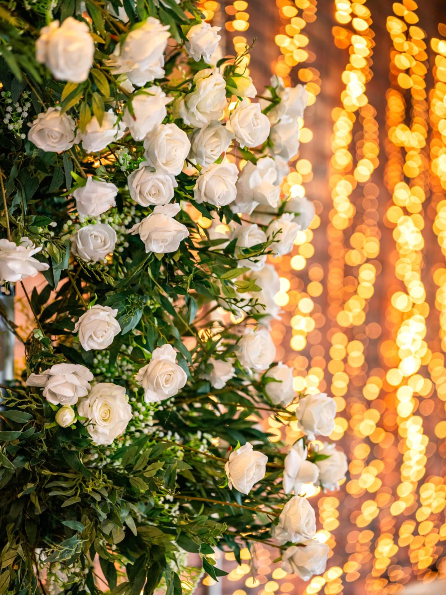 White roses and greenery arranged as elegant flowers for arch wedding decor with warm golden lights in the background.
