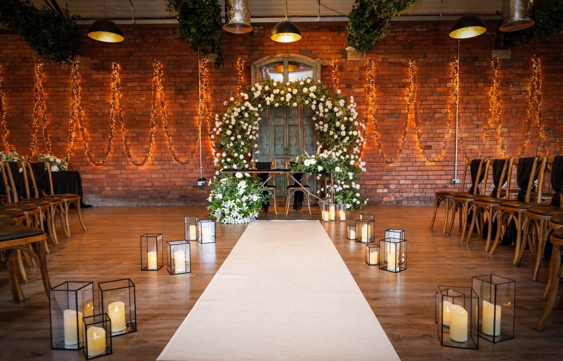 Ceremony setup featuring white arch wedding flowers, greenery, candles and a rustic backdrop with warm fairy lights.