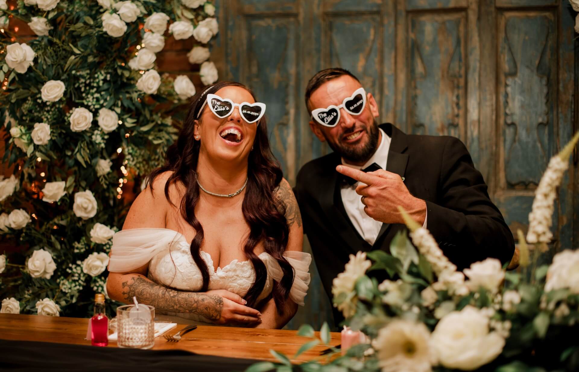 Bride and groom laughing beneath an elegant archway of flowers decorated with white roses and greenery.