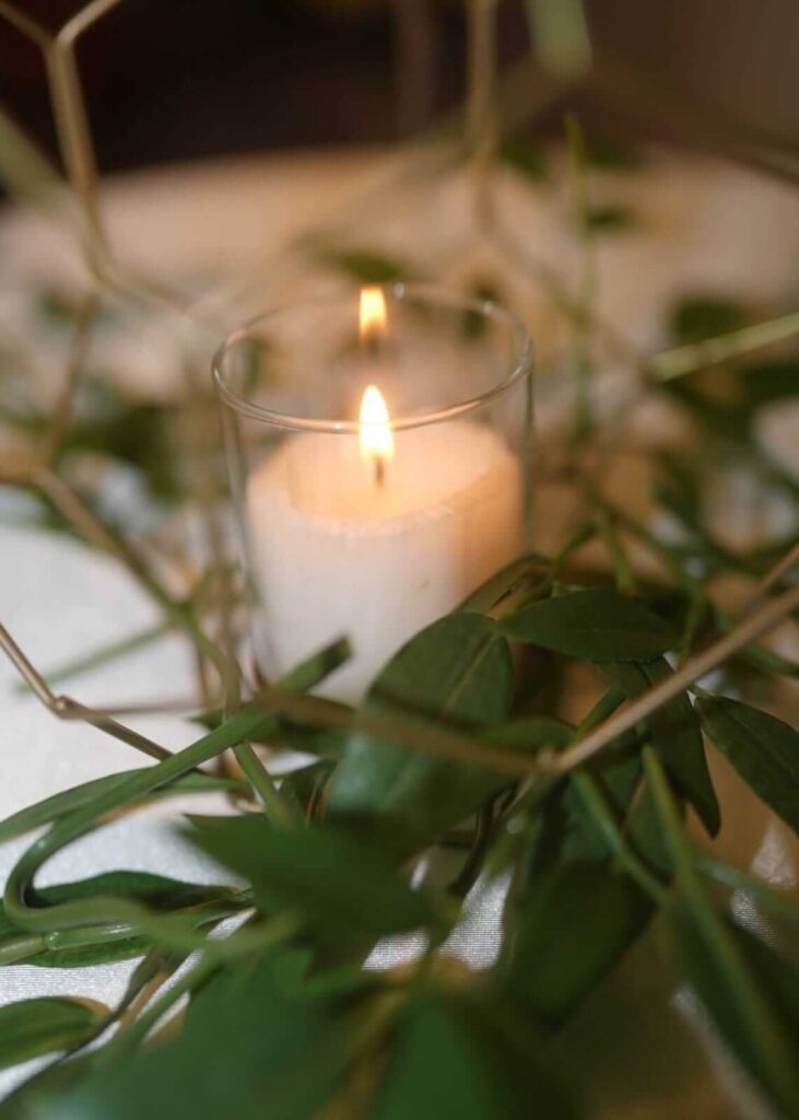 White candle in a glass holder with greenery, styled for popular wedding themes and elegant table decor.