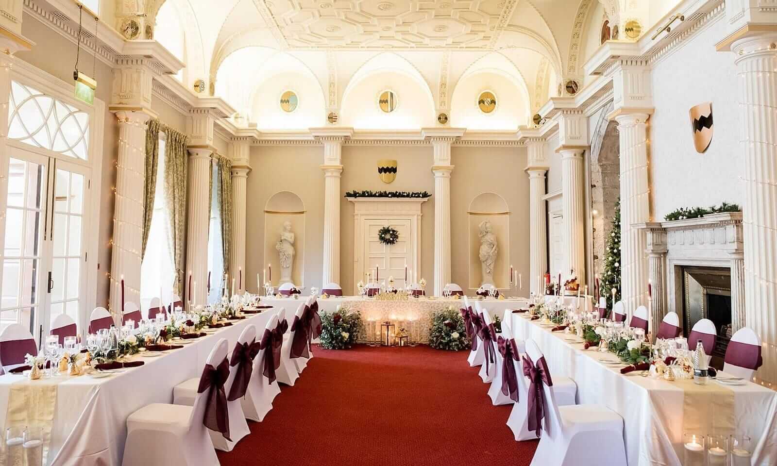 Elegant venue styling with long banquet tables, white chair covers and burgundy bows in a grand wedding hall.