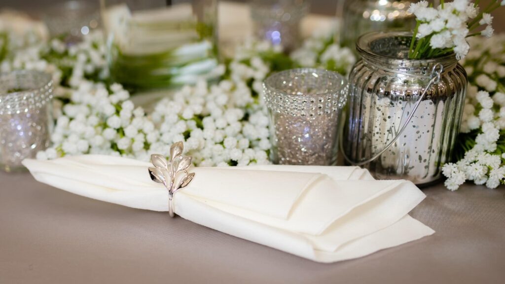 Wedding room table decor with ivory napkin in silver leaf ring, mercury glass votives and baby’s breath garland.