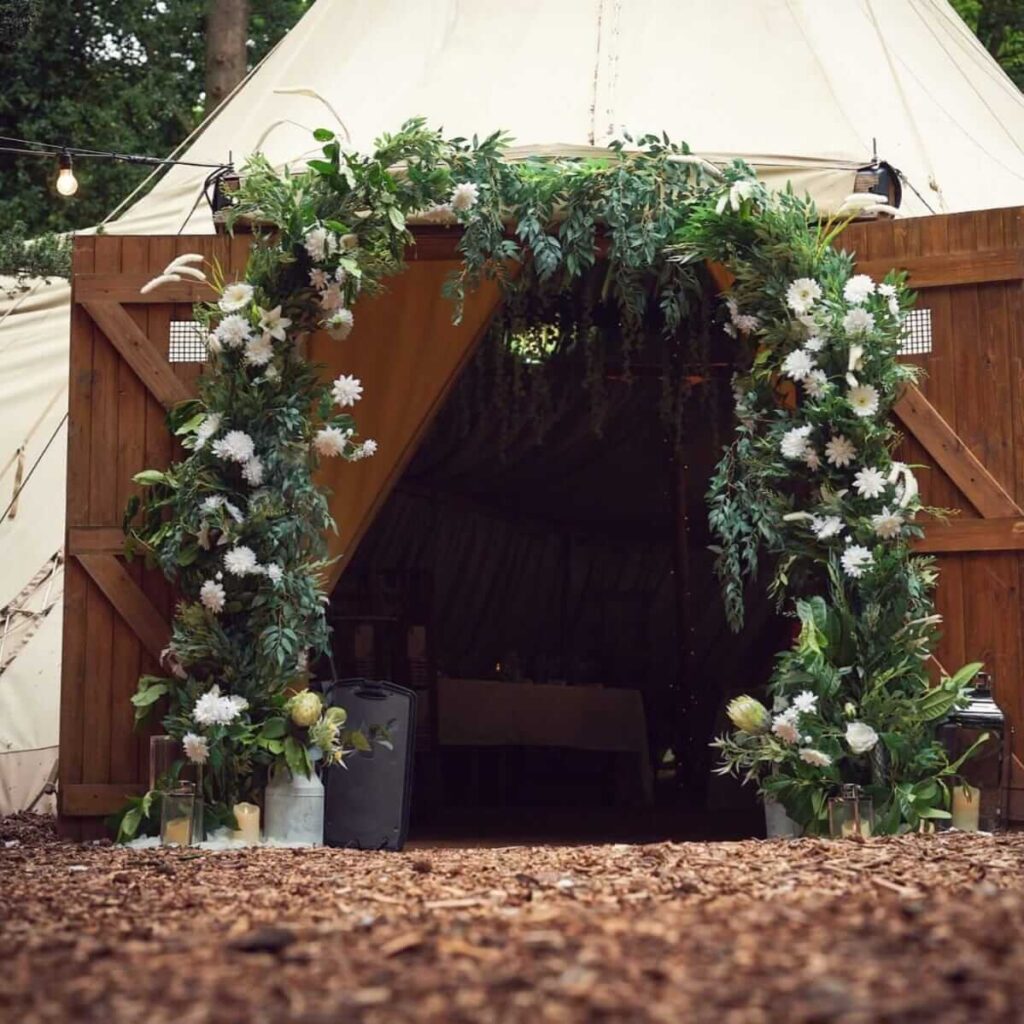 Event dresser floral arch framing tipi entrance with greenery and white flowers.