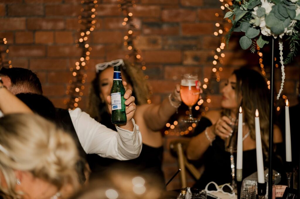 DIY event decor with guests raising drinks at a candlelit table and fairy lights against a brick wall.