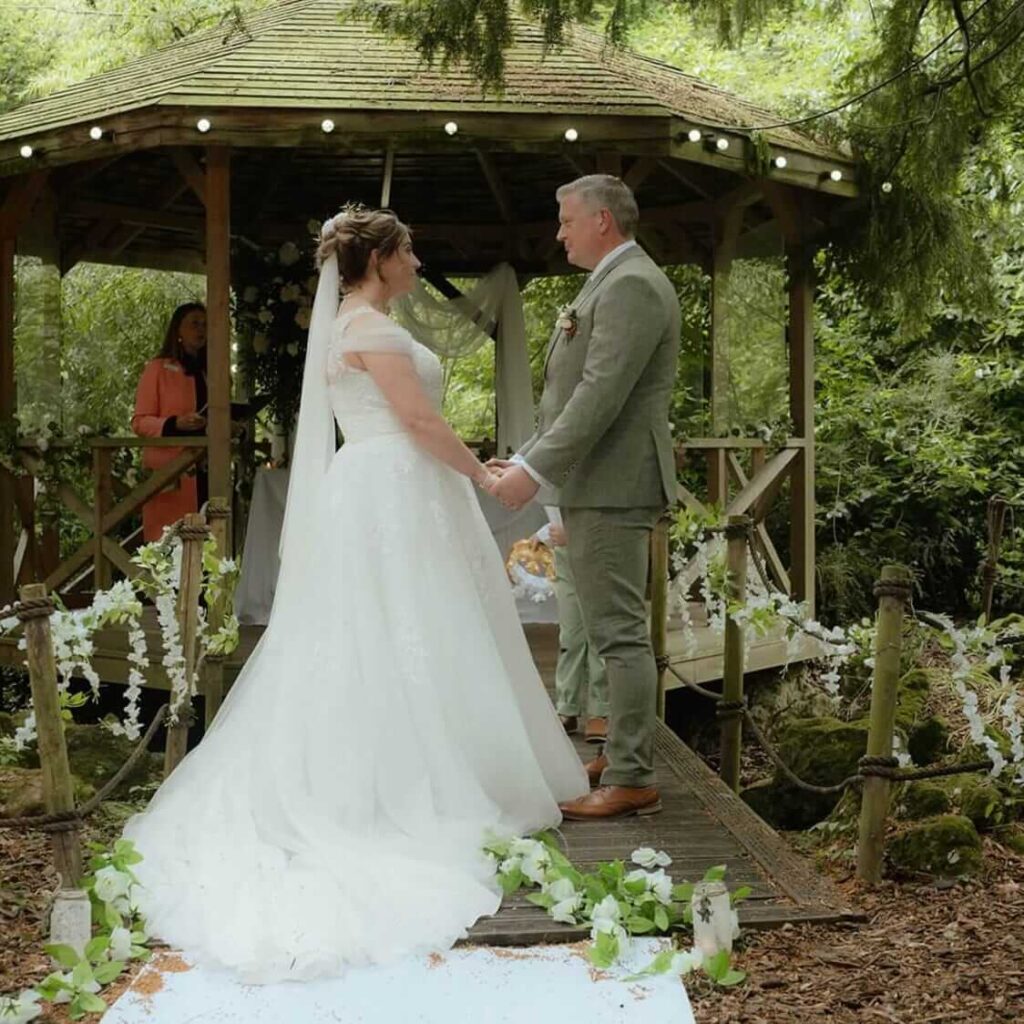 Woodland wedding ceremony with the couple holding hands on a rustic bridge in front of a gazebo.